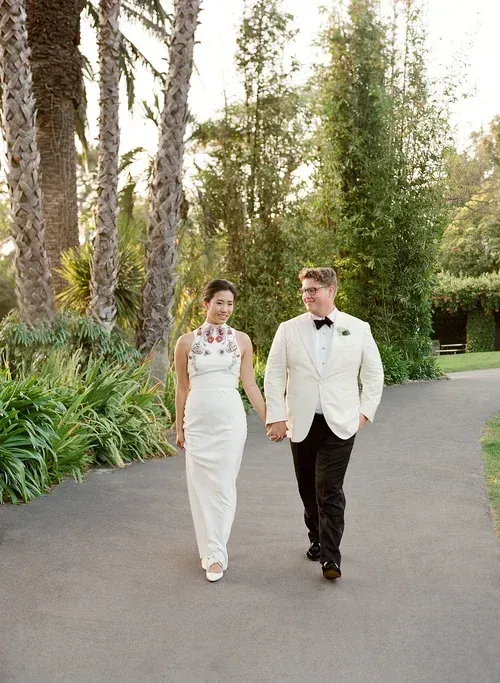 Two people in formal attire walking hand in hand along a garden path.