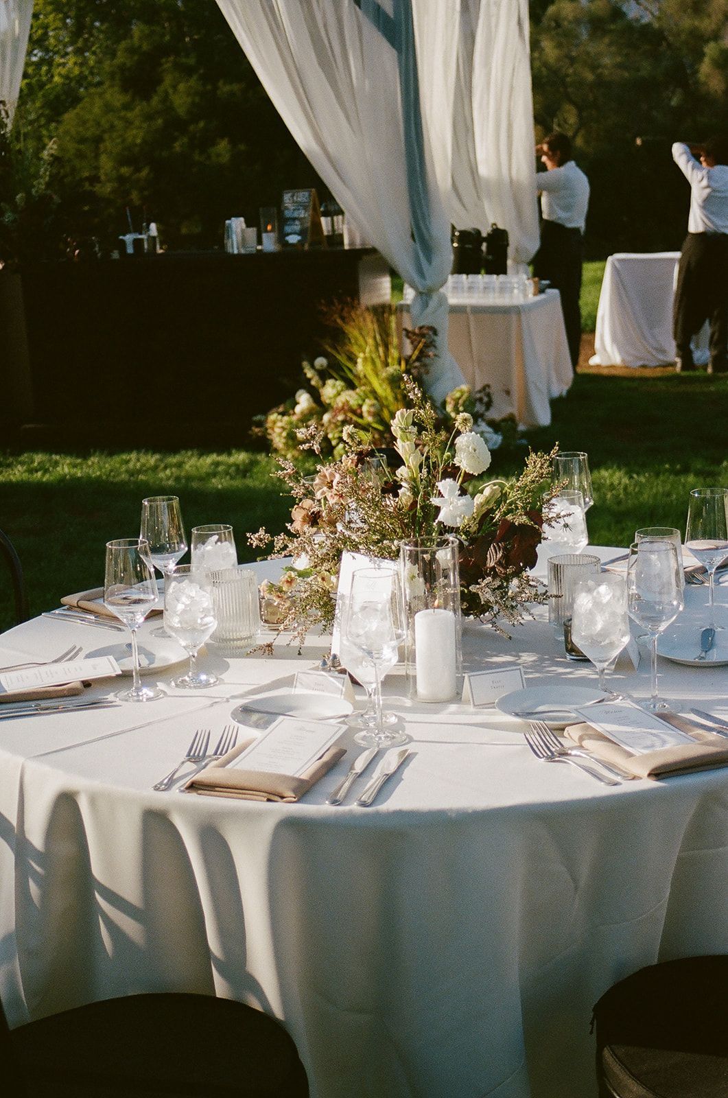 Elegant outdoor dinner table with white linens, floral centerpiece, and place settings under draped canopy.