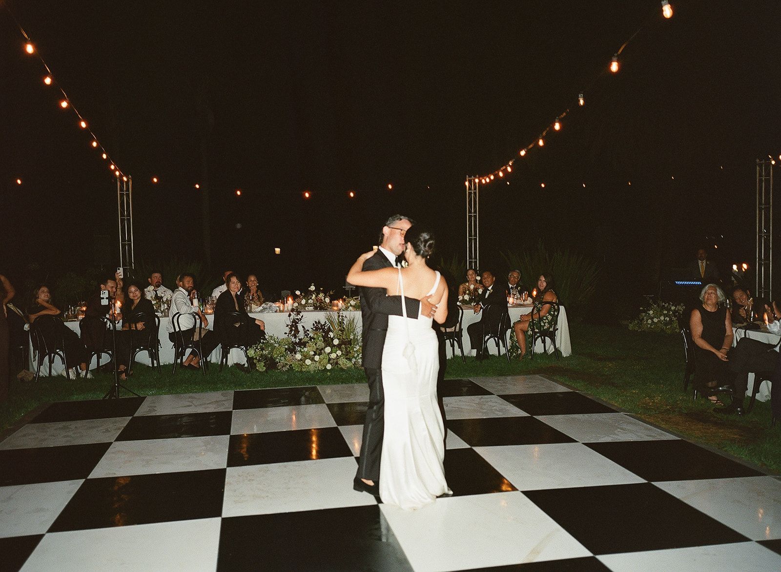 Couple dancing on a black-and-white checkered floor at an outdoor evening reception with string lights
