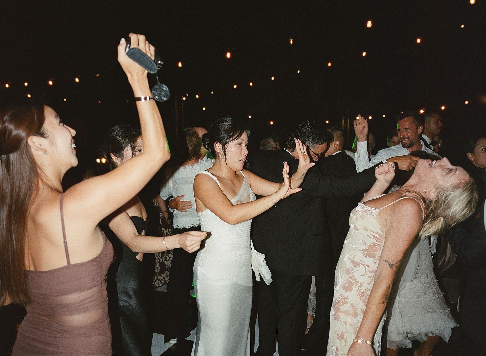 People dancing at a wedding reception under string lights, with a bride in a white dress in the center