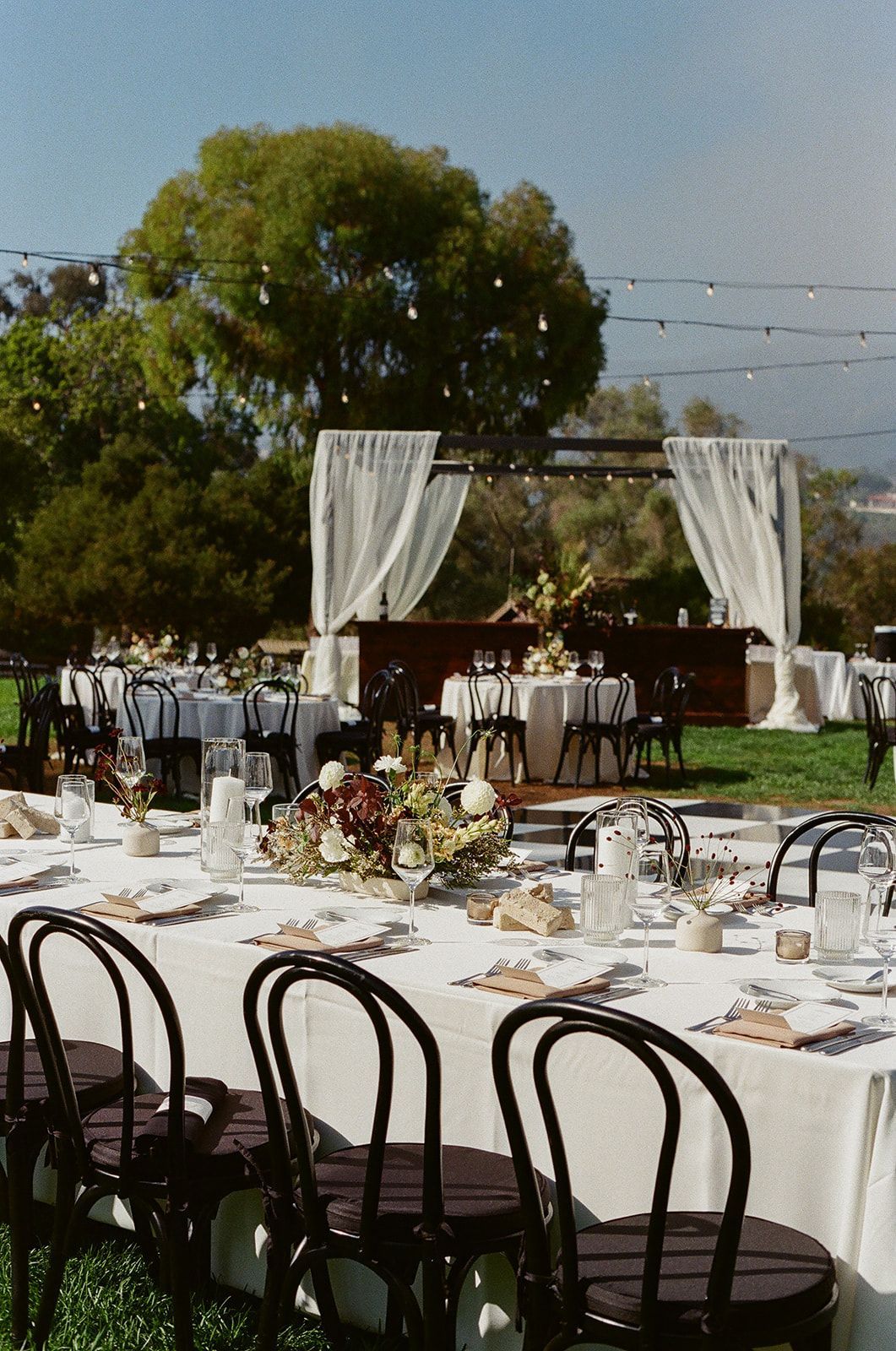 Outdoor wedding reception with round tables, white linens, and a decorated ceremony arch under trees.