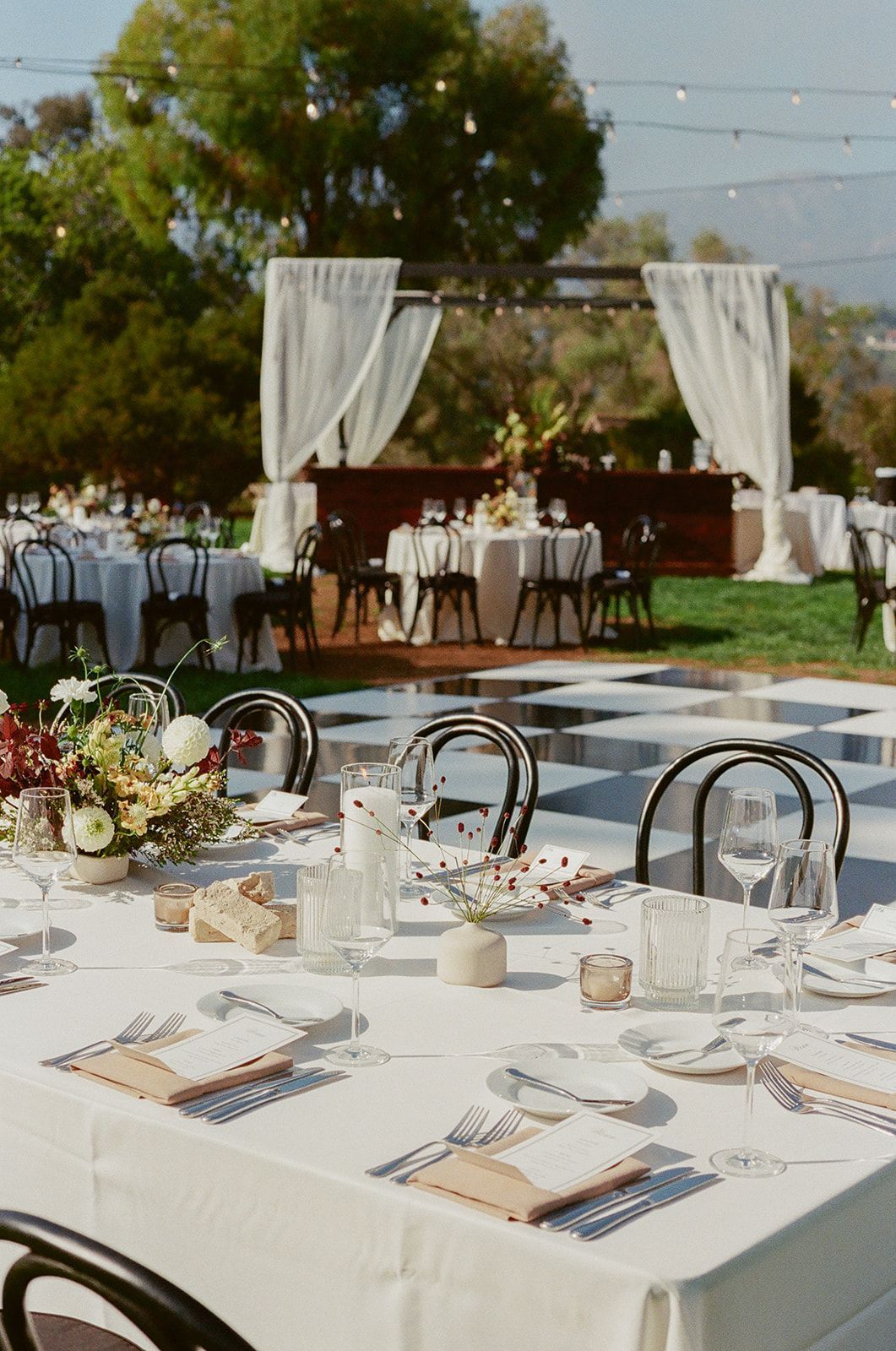 Outdoor wedding reception setup with white table settings, checkered dance floor, and draped arch