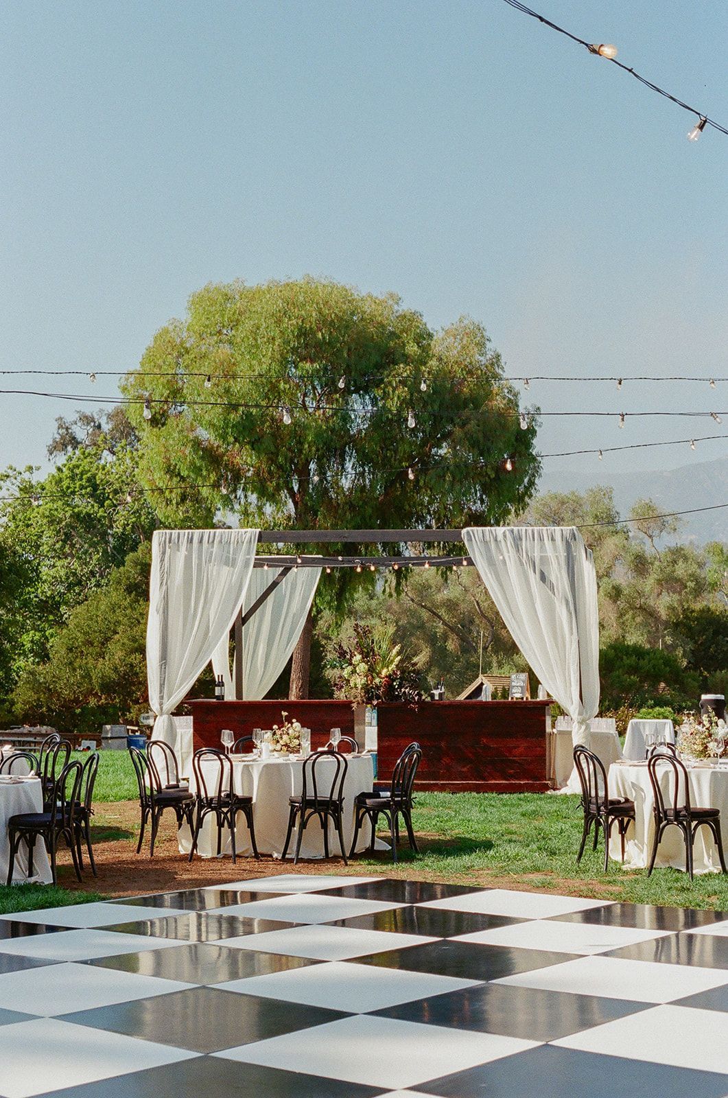Outdoor event setup with a checkered floor, white drapes, and a decorated table under a tree