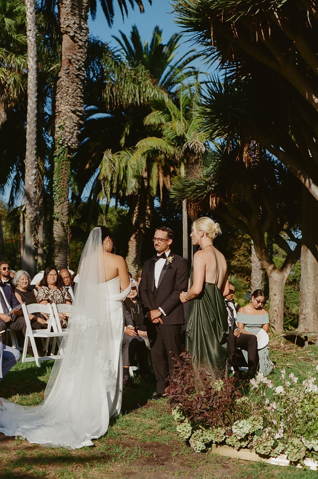 Wedding ceremony outdoors among palm trees, with bride, groom, and officiant standing under bright sunlight.