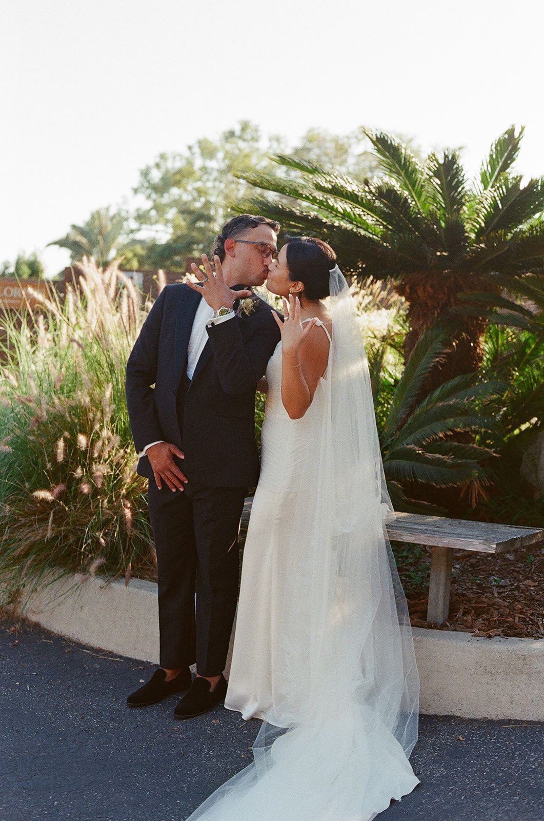 Wedding couple in formal attire kissing outdoors near palm trees and a stone bench.