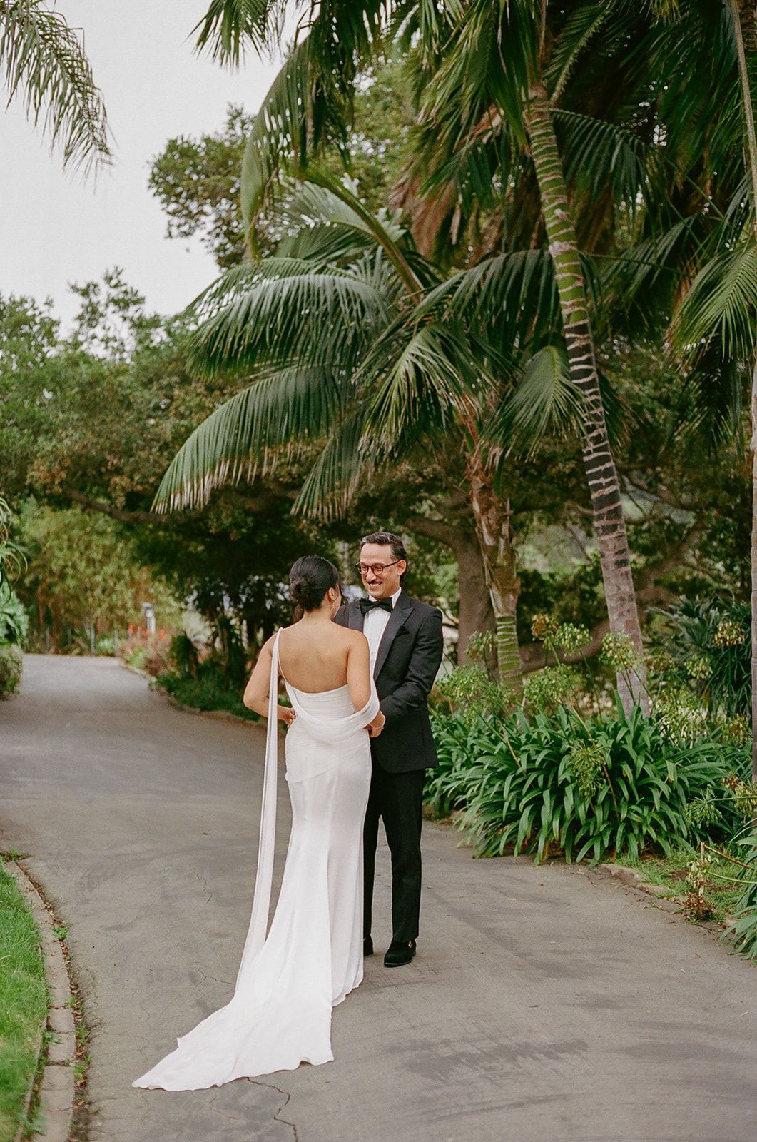 Bride and groom standing on a palm-lined path, facing each other outdoors.