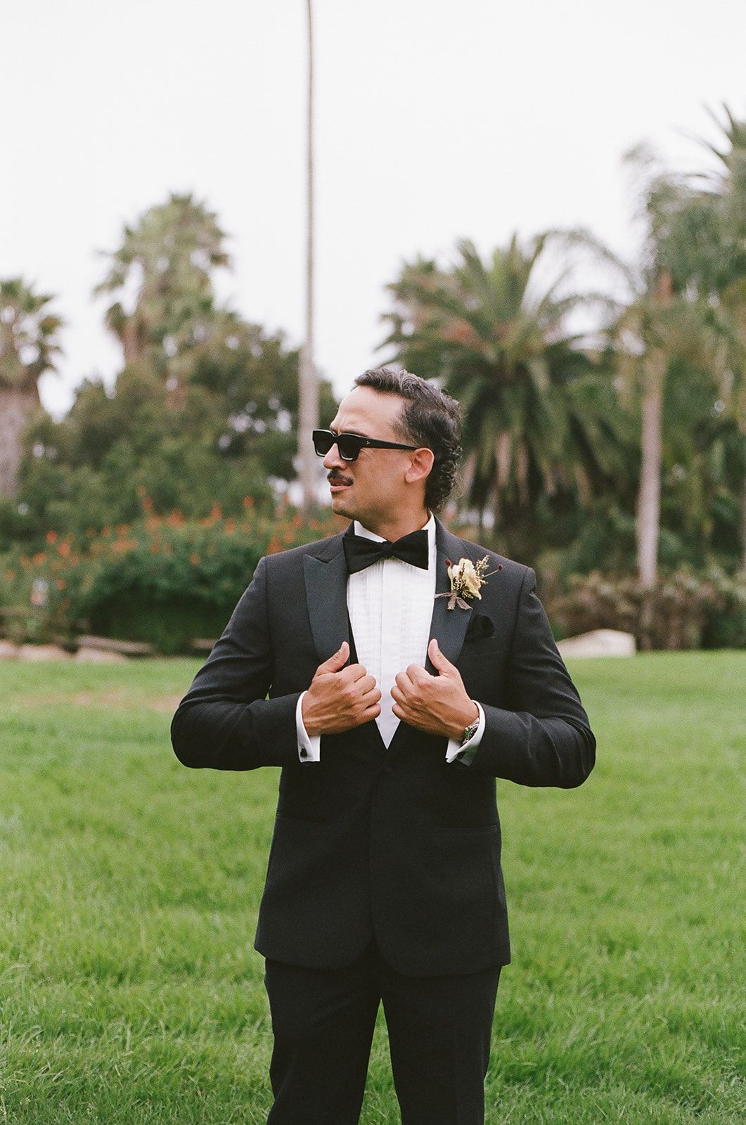 Man in a black tuxedo adjusting his jacket on a grassy lawn with palm trees in the background