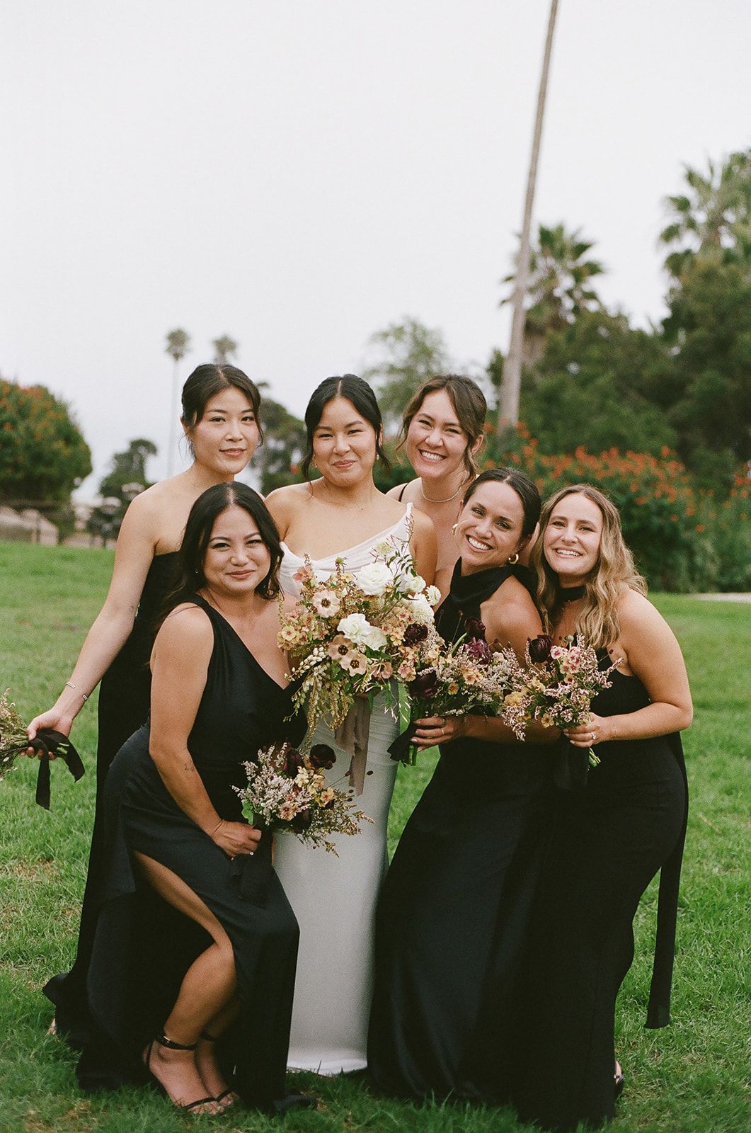 Group posing outdoors in formal dresses, smiling and holding bouquets on a grassy lawn.