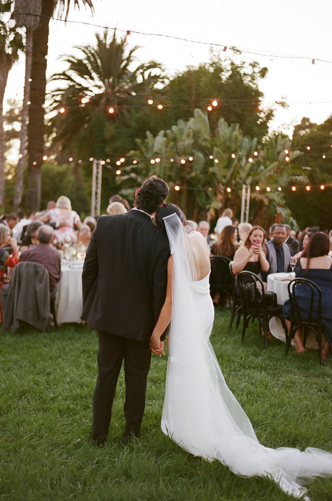 Wedding couple seen from behind walking on a lawn at an outdoor evening reception