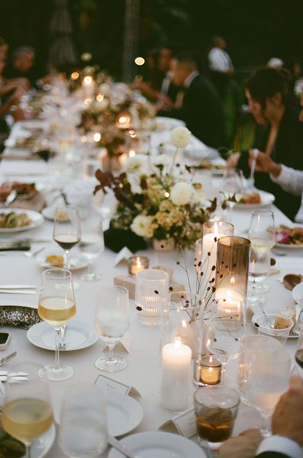 Elegant candlelit dinner table with white linens, floral centerpieces, and wine glasses outdoors at dusk