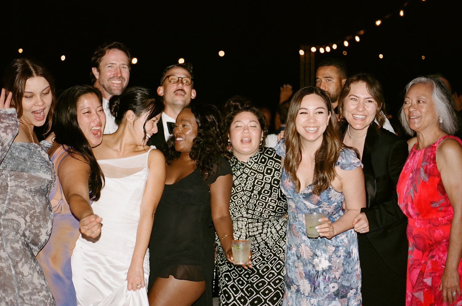 Group of people smiling and dancing at an outdoor nighttime celebration with string lights.