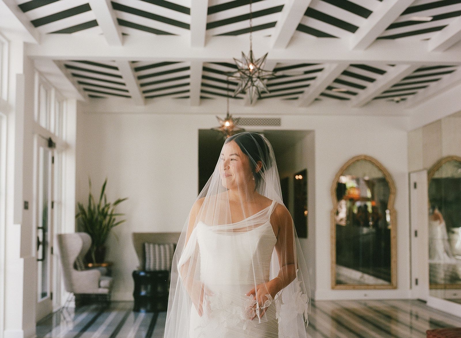 Bride in a white gown and veil standing in a bright hallway with tiled floors and chandeliers