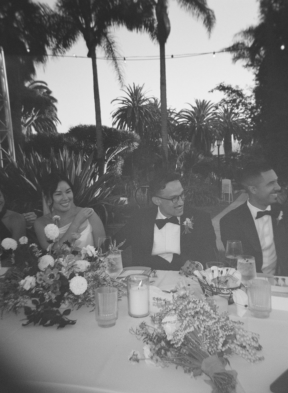 Formal dinner table with floral centerpieces and drinks, three people seated outdoors at night with palm trees behind them