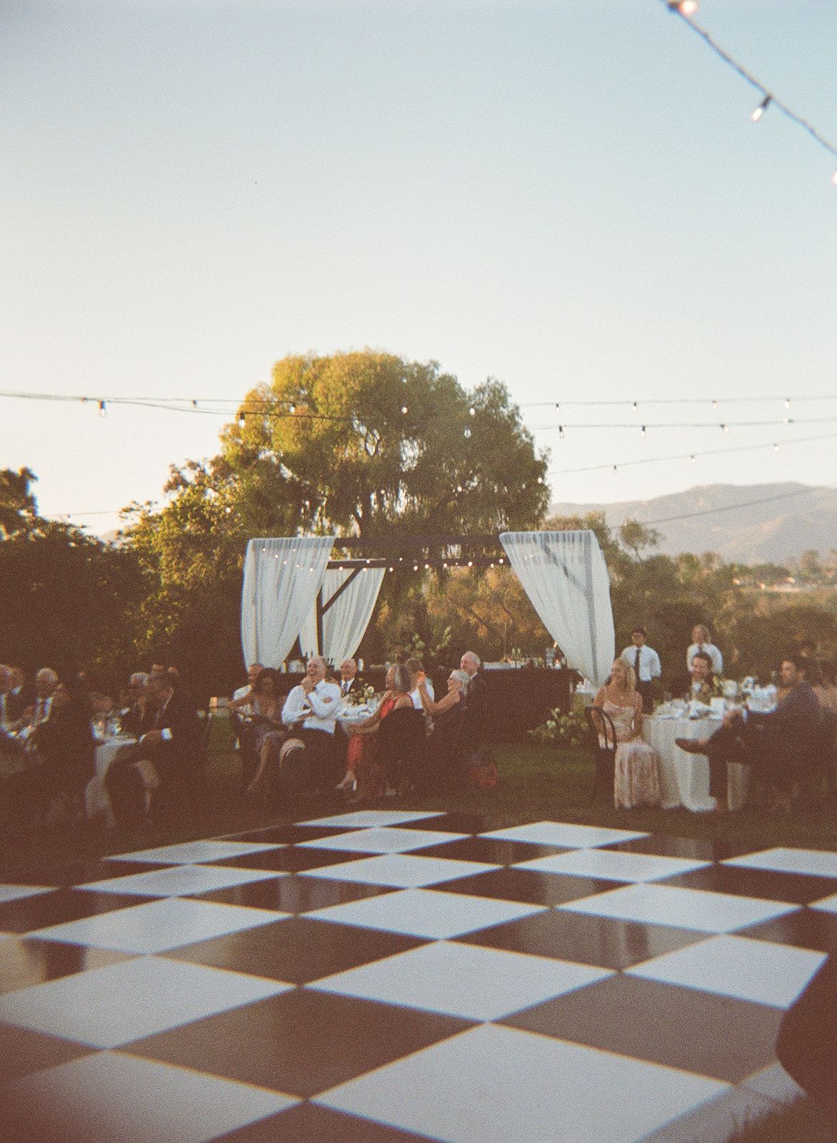 Outdoor gathering beside a black-and-white checkerboard dance floor with guests under string lights and white drapes