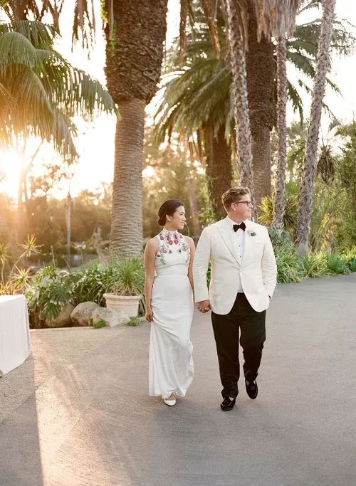 Two people in white formalwear walking hand in hand along a palm-lined path at sunset.