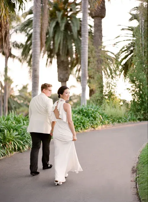 Two people in light formalwear walking on a palm-lined path in a garden