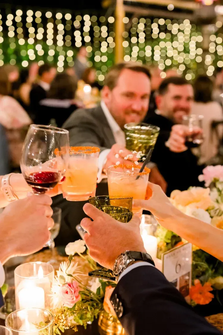 Guests toast with cocktails at a festive outdoor dinner under warm string lights.