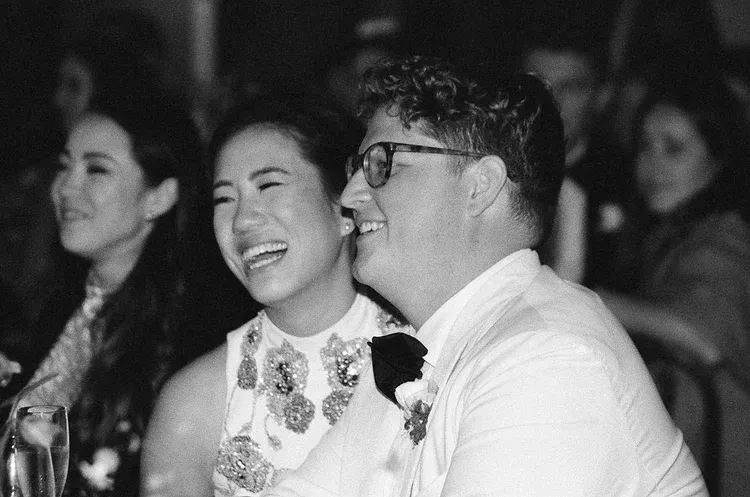 Two people smiling and laughing at a dimly lit event, one in glasses and a white shirt with a boutonniere.