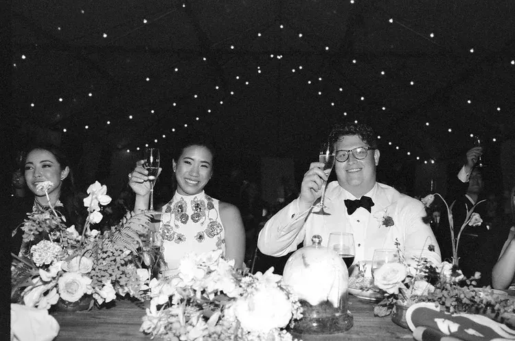 Three people seated at a decorated banquet table, smiling and raising drinks under starry lights.