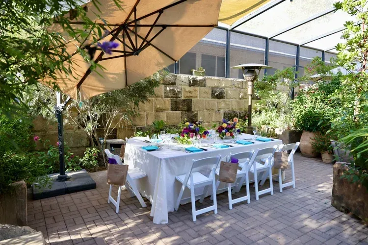Outdoor patio dining table set for a meal under umbrellas, surrounded by lush plants and brick paving.