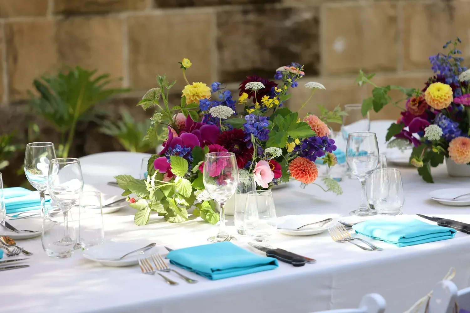 Elegant outdoor table set with white linens, blue napkins, and colorful floral centerpieces