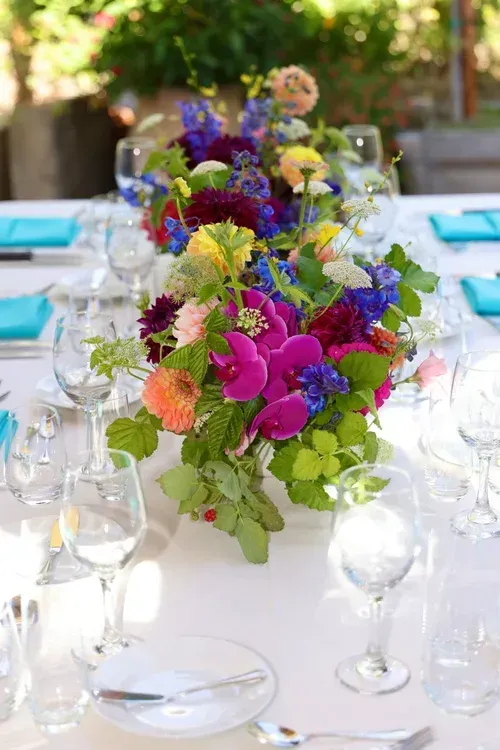 Colorful flower centerpiece on a white dining table with glasses and plates at an outdoor event
