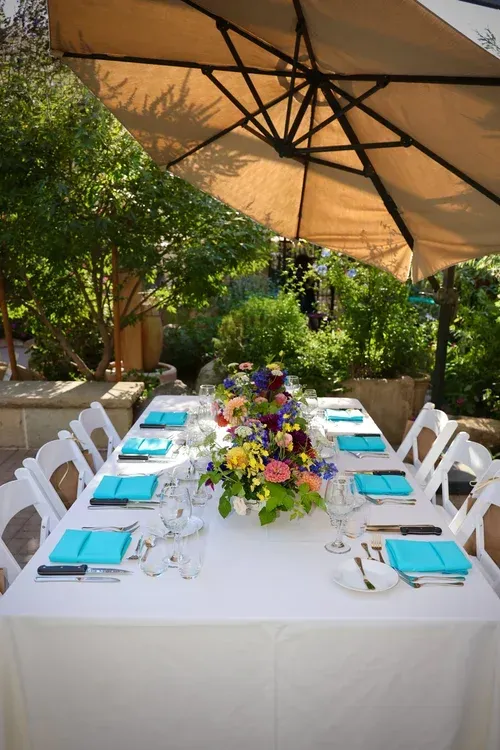 Outdoor dining table with white chairs, turquoise napkins, and a colorful floral centerpiece under a patio umbrella