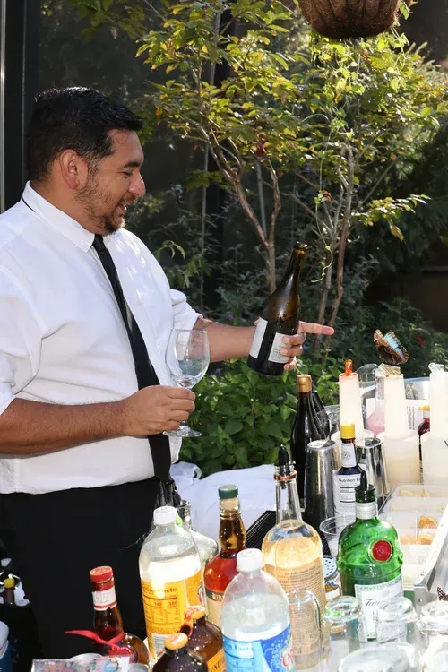 Bartender in white shirt pouring a drink at an outdoor table with assorted liquor bottles and glasses.