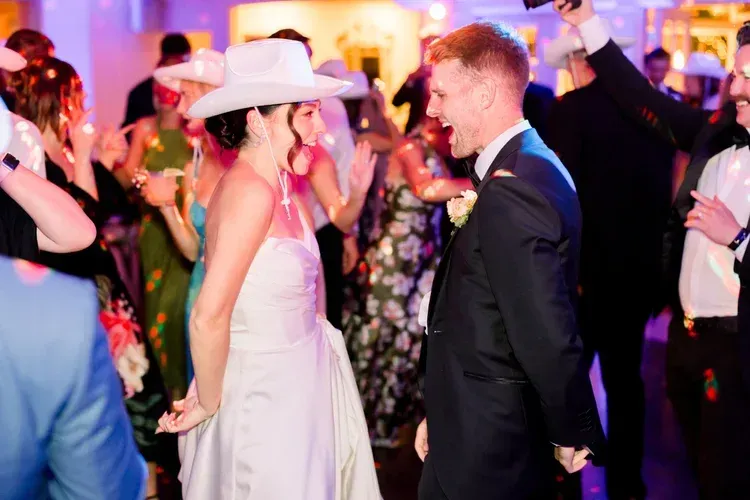 Bride and groom dancing at a lively wedding reception, smiling under warm pink and purple lights