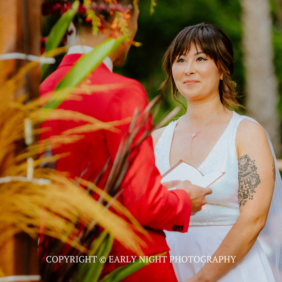 Woman in white dress holding papers, smiling during an outdoor ceremony with a person in red.