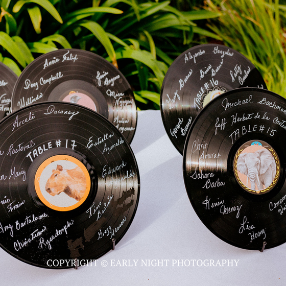 Vinyl record table cards with handwritten guest names on white cloth outdoors, near green plants