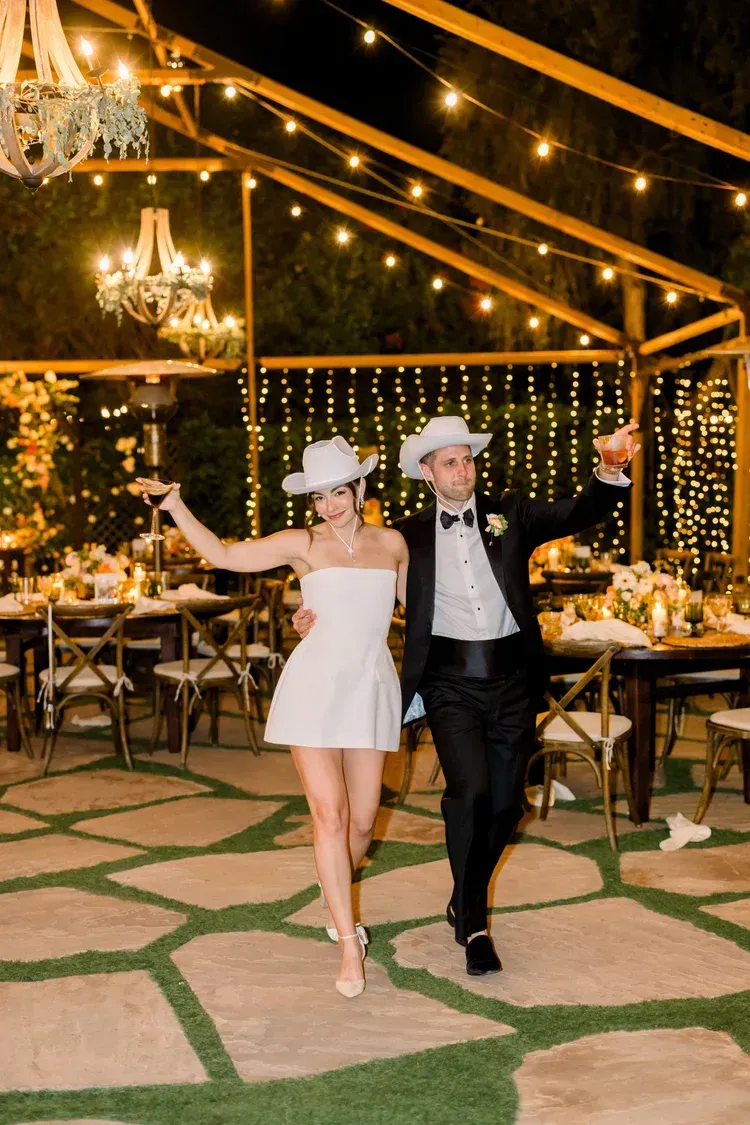 Two people in white hats posing under string lights at an outdoor evening celebration