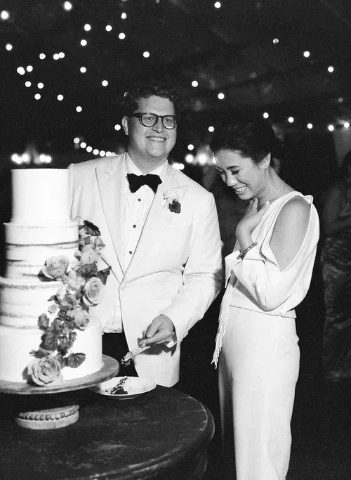 Couple cutting a tiered wedding cake at a decorated reception, both dressed in formal attire.