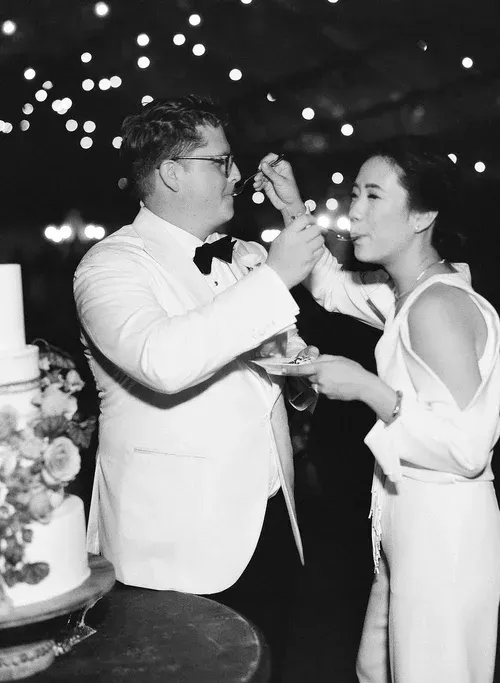 Couple in white formalwear feeding each other cake at a nighttime celebration with twinkling lights