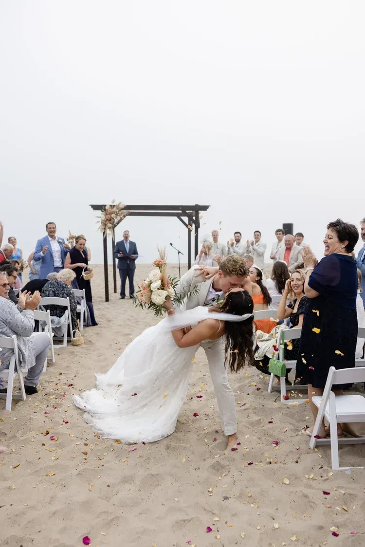 Bride and groom kiss in a beach wedding aisle lined with seated guests and white chairs