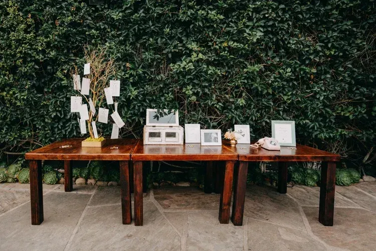 Long wooden table with decorative items and small framed displays against a green hedge backdrop