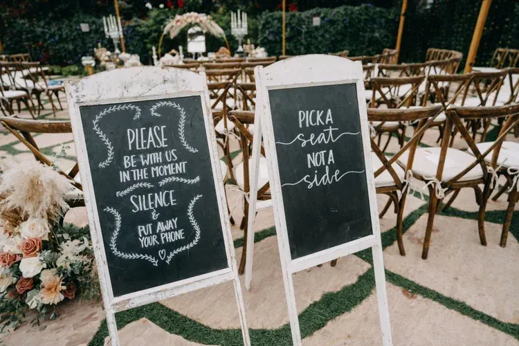 Wooden chalkboard signs at an outdoor wedding read “Please be sure to sign in the guest book” and “Pick a seat, not a side.”