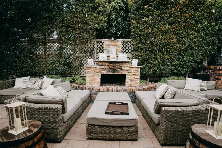 Outdoor patio seating with wicker sofas around a fire pit and stone fireplace, backed by dense greenery
