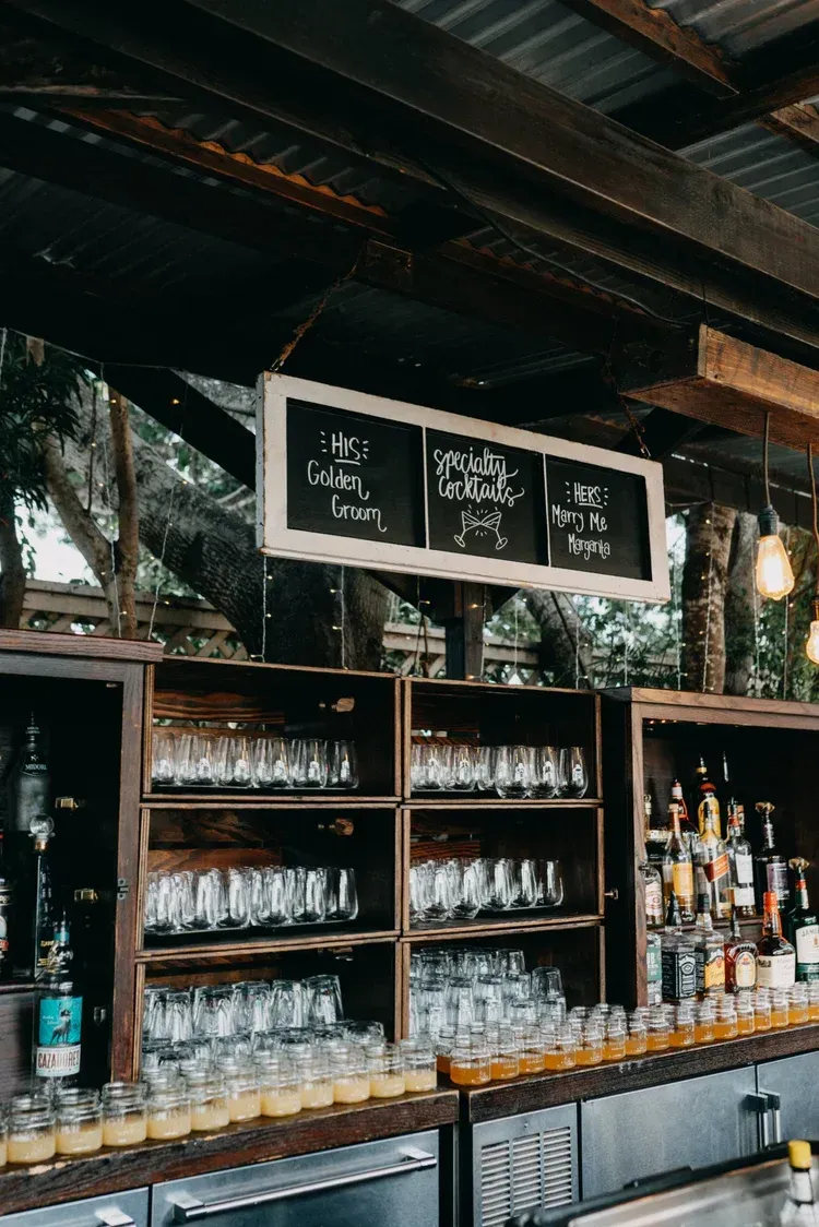 Rustic outdoor bar with liquor bottles, glassware, and chalkboard menus under a wooden roof