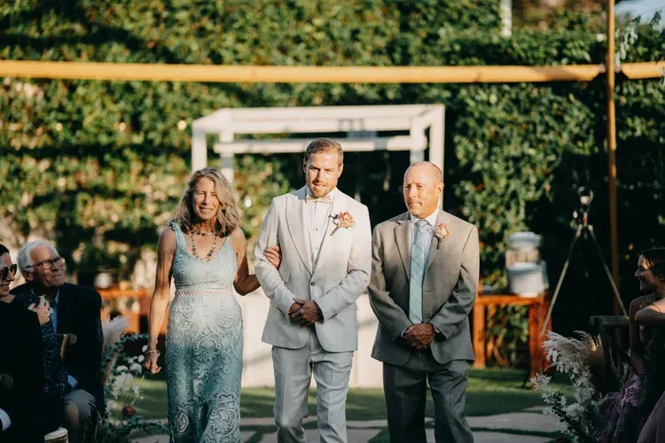 Three people in formal attire walk down an outdoor aisle at a wedding ceremony.