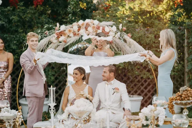 Outdoor wedding ceremony with bride and groom seated beneath a floral arch, attendants holding draped fabric