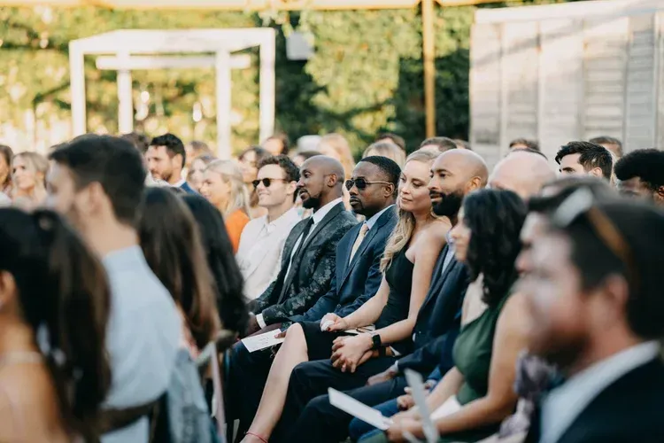People seated outdoors at a formal event, facing forward in a sunny garden setting.