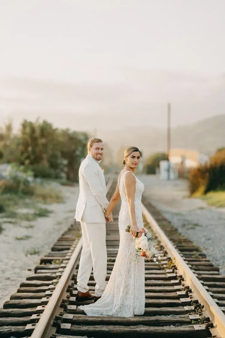 Bride and groom standing on railroad tracks in white wedding attire at sunset