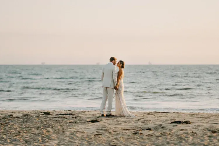 Couple in white outfits standing on a beach, facing the ocean at sunset.