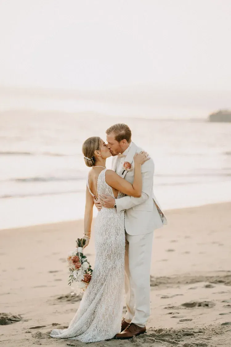 Bride and groom kissing on a beach at sunset, bride holding a bouquet