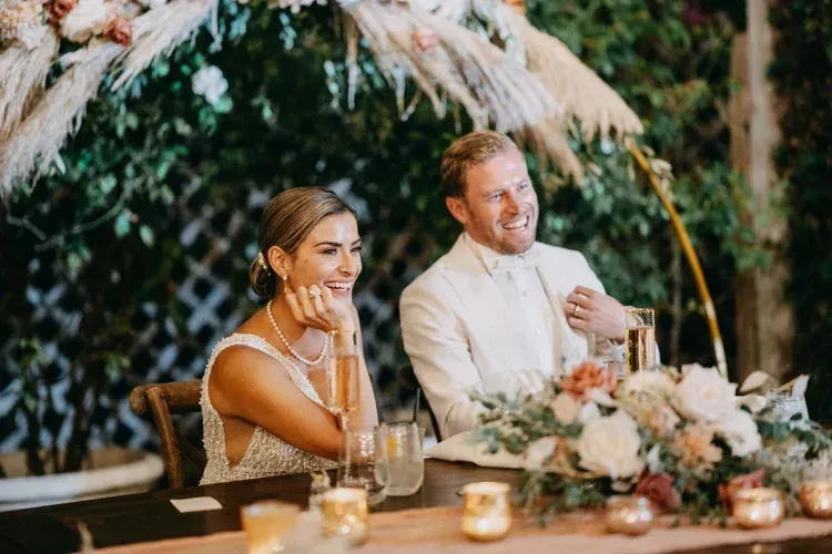 Couple seated at a decorated dinner table in a lush outdoor setting, smiling during a celebration