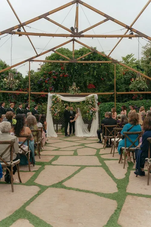 Wedding ceremony under a rustic open-air arch with guests seated on both sides of a stone aisle