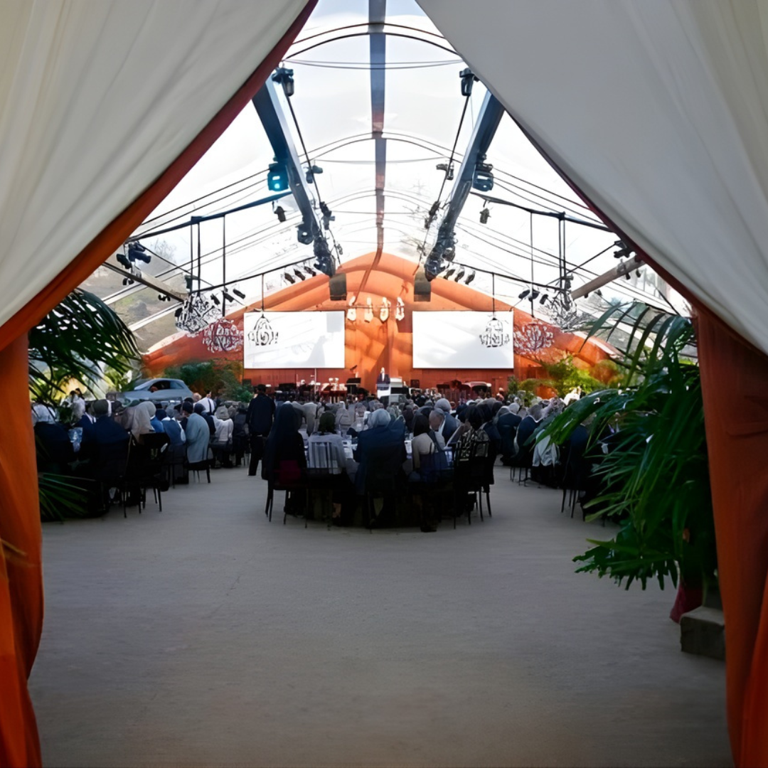Crowded indoor event under white and orange draped tent, with stage and screens at the far end