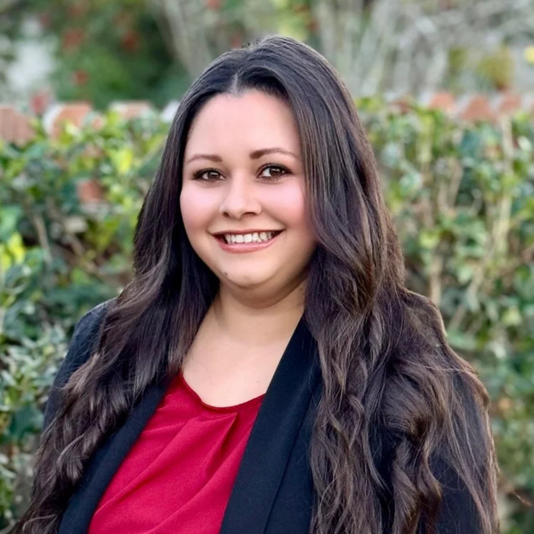 Smiling person with long dark hair in a red top standing outdoors in front of greenery