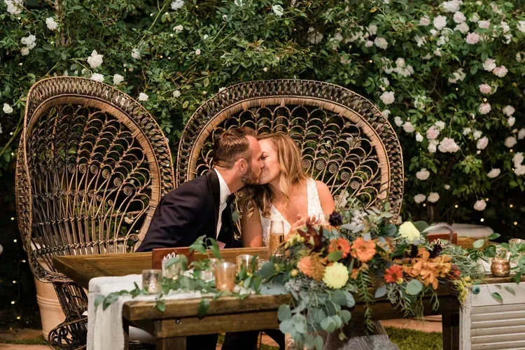 Wedding couple kissing at a flower-covered table with wicker chairs and floral arrangements